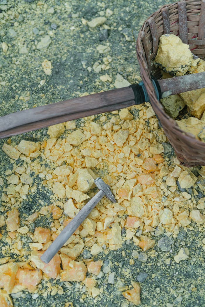 Close-up of a hammer and basket amidst yellow sulfur rocks on the ground.